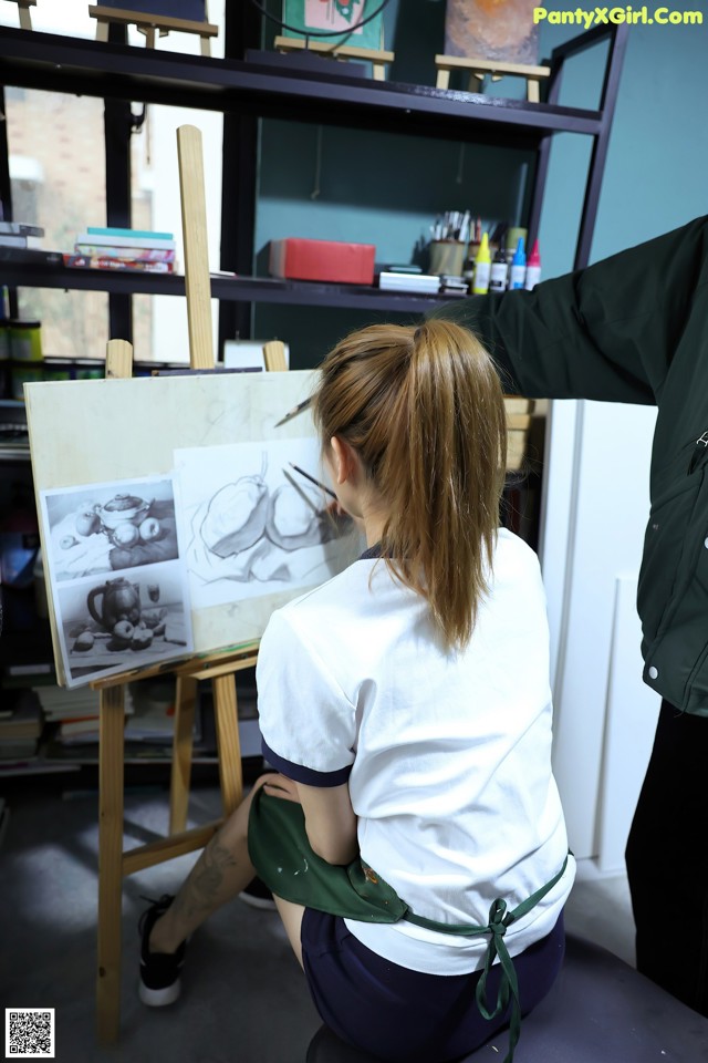 A woman sitting on a stool in front of an easel painting.