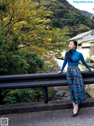 A woman in a blue sweater and a plaid skirt is standing by a window.