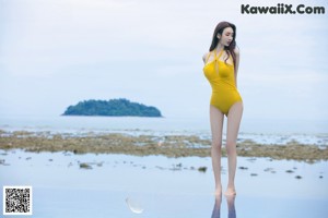 A woman in a white bathing suit leaning against a tree on the beach.